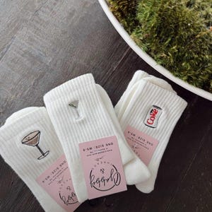 three pairs of white grip socks with different embroidered icons. One has an espresso martini, one has a dirty martini, diet coke