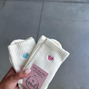 Two pairs of White socks with an embroidered heart held by a hand against a gray background