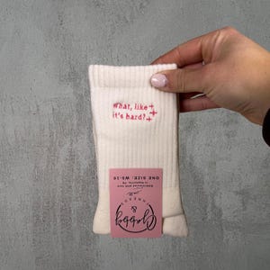 a pair of white socks with the text "what like its hard" embroidered on it against a white background