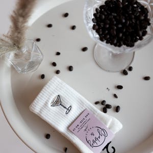 a pair of white socks with an embroidered espresso martini sits on a table with espresso beans