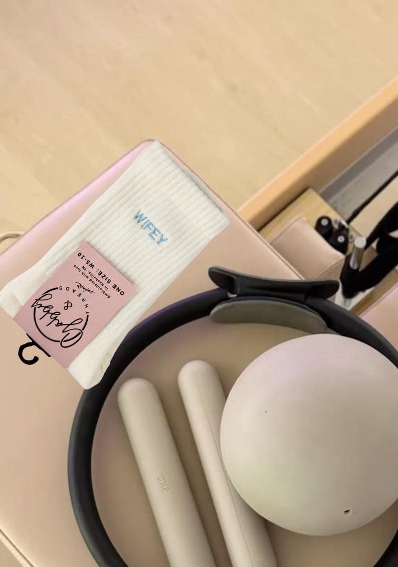 a pair of white grip socks with the word "wifey" embroidered on them sits next to workout equipment on a reformer