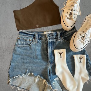 a pair of denim shorts, a brown sports bra, white converse and embroidered espresso martini socks against a grey background