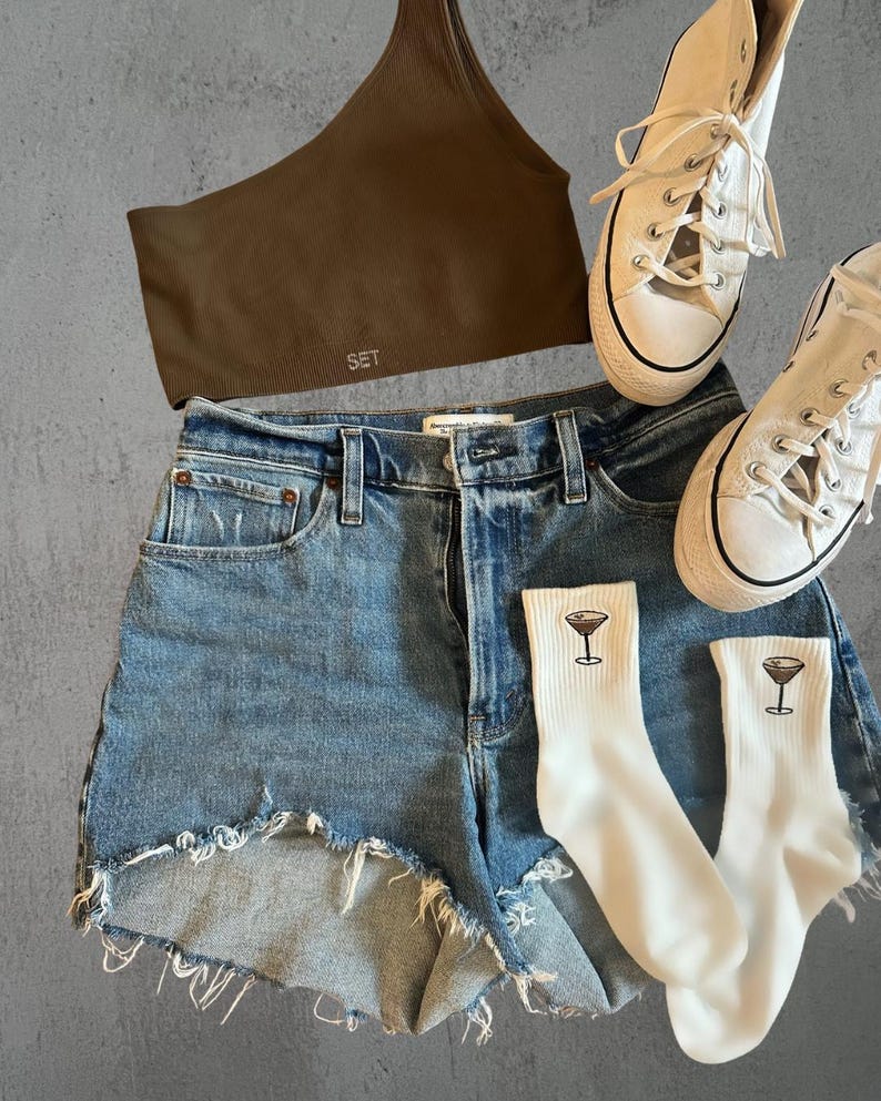 a pair of denim shorts, a brown sports bra, white converse and embroidered espresso martini socks against a grey background