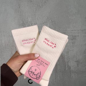 two pairs of white socks with the text "what like its hard" embroidered on them against a white background