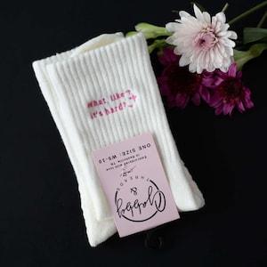 a pair of white grip socks with the text "what like it's hard" embroidered on them next to florals