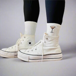 A person is wearing white converse with crew socks that have an embroidered espresso martini detail on them