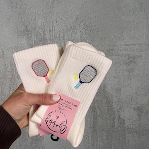 May include: Cream ribbed socks featuring embroidered pickleball paddles and balls. One sock has a red paddle, the other a blue paddle. A pink tag with the text "Gabby & Threads" is attached.