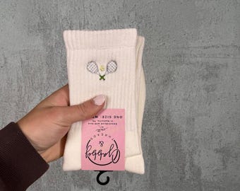 Könnte beinhalten: Cremefarbene gerippte Socken mit einem gestickten Tennis-Schläger- und Ball-Motiv. Die Socken haben ein rosa Etikett mit dem Text "Crabby & Threads" und einen schwarzen Hänger. Einheitsgröße.