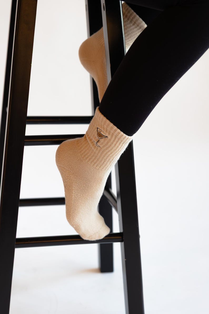 Person wearing beige socks with an embroidered espresso martini n a black stool against a white background
