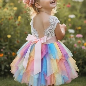 May include: A little girl in a colorful rainbow tutu dress with a pink bow. The dress has a white lace top and a layered skirt in shades of pink, yellow, blue, and purple. A pink flower adorns her hair.