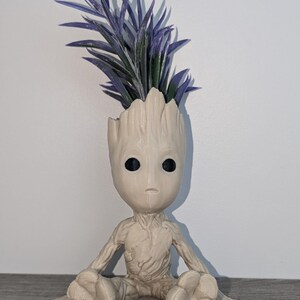 May include: A beige ceramic planter shaped like a sitting Groot character from the Guardians of the Galaxy movie. The planter has a purple and green artificial plant growing out of the top of its head.