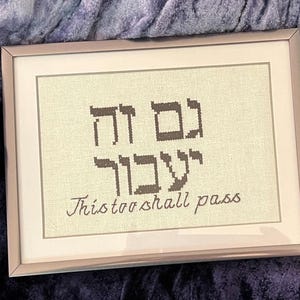 May include: A framed cross-stitch with Hebrew text and the phrase "This too shall pass".
