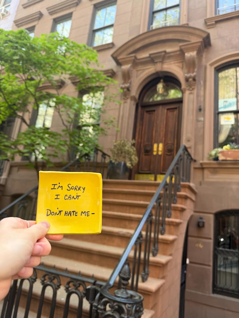 May include: A yellow square ceramic dish with the handwritten text "I'M SORRY I CAN'T DON'T HATE ME-" is held in front of a brownstone building with a wooden door and black railing. The building has multiple windows and a tree.