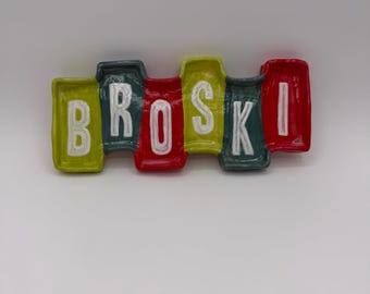May include: A colorful ceramic wall hanging with the word "BROSKI" spelled out in individual, rectangular blocks. The blocks are in shades of yellow, red, green, and white, with a glossy finish. The letters are white.