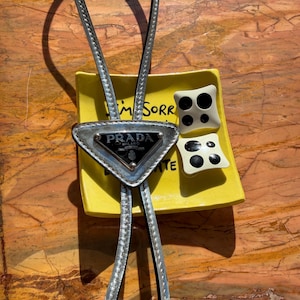 May include: A silver Prada bolo tie with a triangular pendant and the brand's logo. The tie rests on a yellow dish with the words "I'm Sorry" and two decorative black and white square objects.