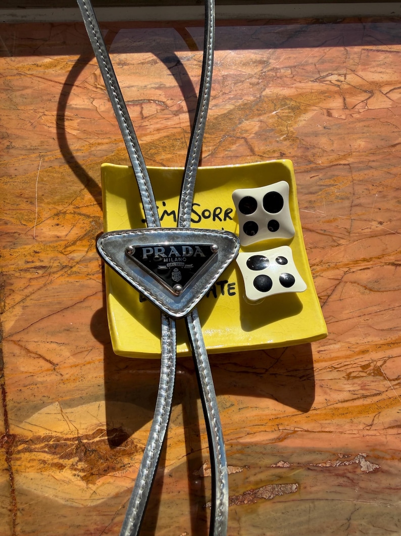 May include: A silver Prada bolo tie with a triangular pendant and the brand's logo. The tie rests on a yellow dish with the words "I'm Sorry" and two decorative black and white square objects.