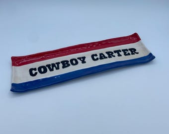 May include: A rectangular ceramic tray with a patriotic color scheme. The top edge is red, the bottom edge is blue, and the center is white with the words "COWBOY CARTER" in black. The tray is slightly curved.