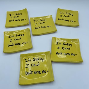 May include: Six yellow ceramic dishes, each with the handwritten text "I'M SORRY I CAN'T DON'T HATE ME-". The dishes are square with slightly rounded edges. The text is in black ink.