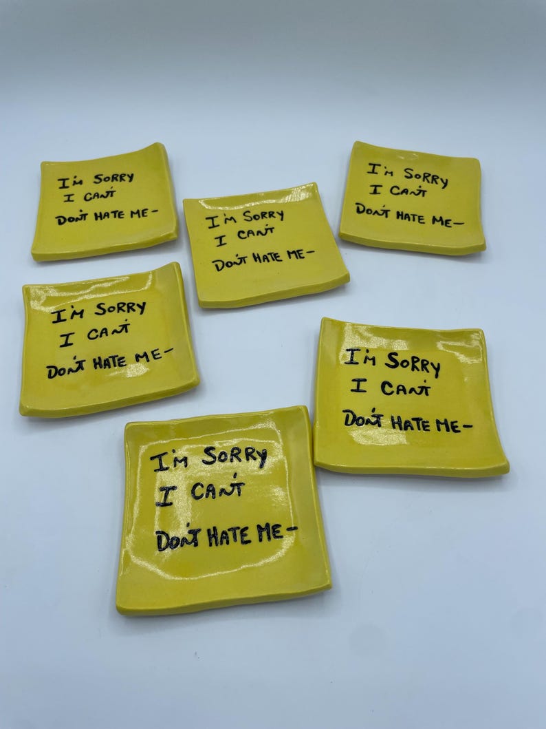 May include: Six yellow ceramic dishes, each with the handwritten text "I'M SORRY I CAN'T DON'T HATE ME-". The dishes are square with slightly rounded edges. The text is in black ink.