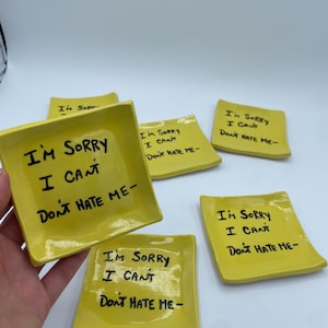 May include: A collection of small, square, yellow ceramic dishes. Each dish has the handwritten phrase "I'M SORRY I CAN'T DON'T HATE ME-" in black ink. The dishes are arranged on a white surface.