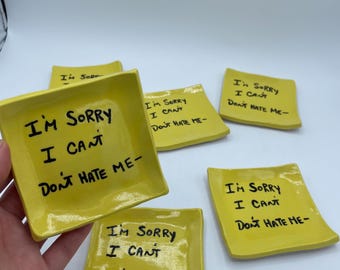May include: A collection of small, square, yellow ceramic dishes. Each dish has the handwritten phrase "I'M SORRY I CAN'T DON'T HATE ME-" in black ink. The dishes are arranged on a white surface.