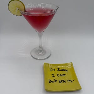 May include: A pink cocktail in a stemmed glass with a lime garnish. A small, yellow ceramic dish with the handwritten text "I'M SORRY I CAN'T DON'T HATE ME" sits in front of the glass. The drink is set against a white background.