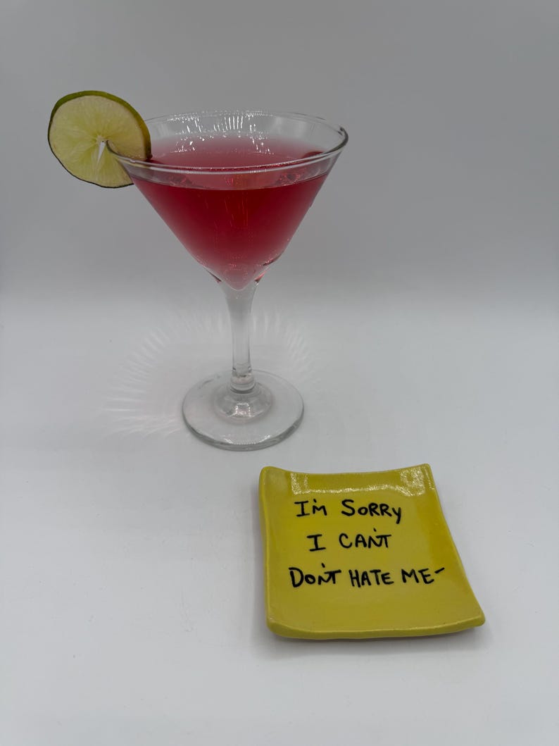 May include: A pink cocktail in a stemmed glass with a lime garnish. A small, yellow ceramic dish with the handwritten text "I'M SORRY I CAN'T DON'T HATE ME" sits in front of the glass. The drink is set against a white background.