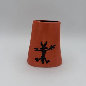 May include: A terracotta-colored ceramic vase with a black interior and a black cartoon coyote design on the front. The vase has a trapezoidal shape and is likely handmade.