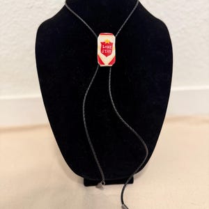 May include: A bolo tie featuring a rectangular pendant shaped like a beer can with a white, red, and yellow Lone Star logo. The pendant is attached to a thin, black braided cord that ends in silver-toned metal tips at the bottom of the strands.