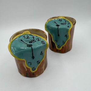 May include: Two ceramic cups with a melting clock design. The clocks have a teal face with black hands and yellow trim. The cups are brown with a textured appearance, resembling wood.