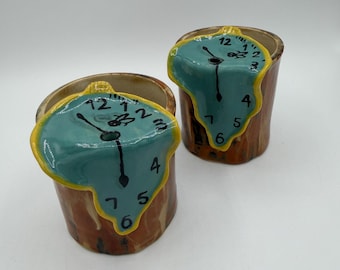 May include: Two ceramic cups with a melting clock design. The clocks have a teal face with black hands and yellow trim. The cups are brown with a textured appearance, resembling wood.