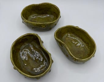 May include: Three handmade ceramic bowls in varying sizes. The bowls have an organic, irregular shape and a glossy, olive green glaze. Each bowl features small, textured bumps around the rim, adding a unique tactile element. The bowls are suitable for serving or display.