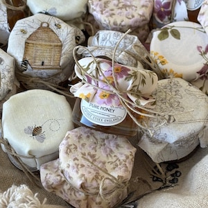 May include: A collection of small glass jars filled with honey, each wrapped in a floral patterned fabric and tied with twine. The jars are arranged in a basket and feature a label that reads "Pure Honey" with the location "Wipptree, Essex, England".