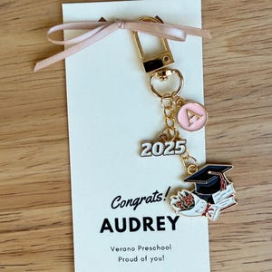 K&ouml;nnte beinhalten: Ein goldfarbener Schl&uuml;sselanh&auml;nger mit einem rosa Initialen-Anh&auml;nger, der Zahl "2025" und einem Abschlusskappen-Anh&auml;nger. Der Schl&uuml;sselanh&auml;nger ist an einer Karte mit dem Text "Congrats! AUDREY" und einem Band befestigt.