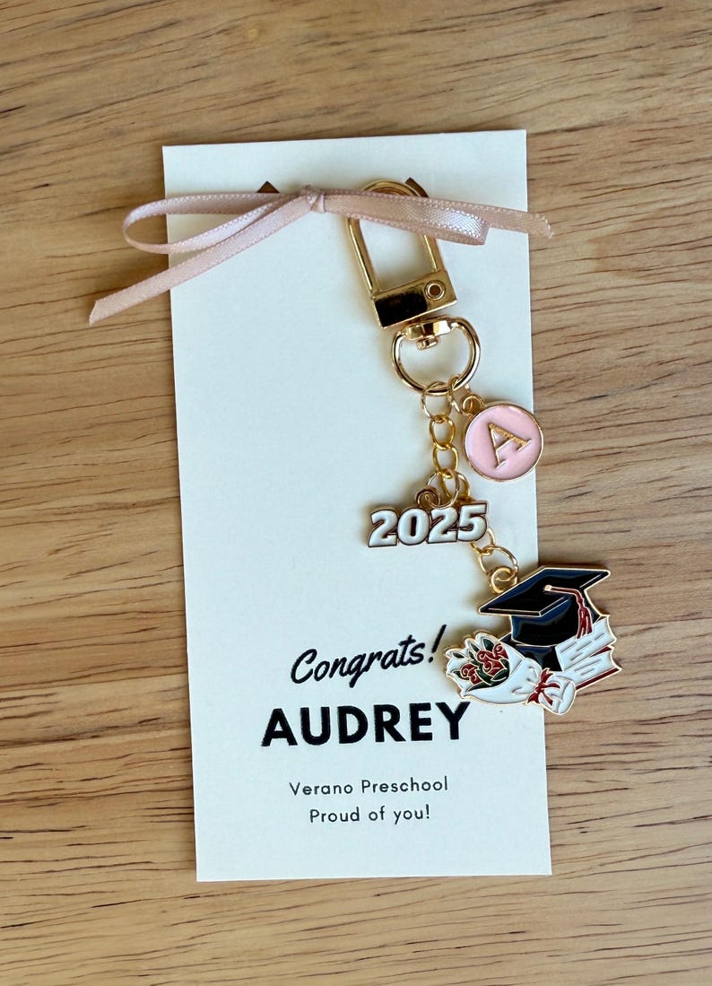 K&ouml;nnte beinhalten: Ein goldfarbener Schl&uuml;sselanh&auml;nger mit einem rosa Initialen-Anh&auml;nger, der Zahl "2025" und einem Abschlusskappen-Anh&auml;nger. Der Schl&uuml;sselanh&auml;nger ist an einer Karte mit dem Text "Congrats! AUDREY" und einem Band befestigt.