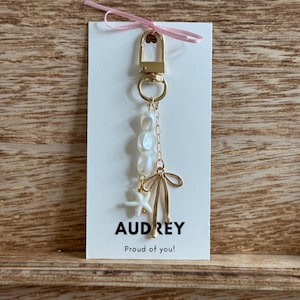 May include: A gold-colored keychain with a white card. The keychain features a lobster clasp, a ring, a chain, and decorative elements including pearls, a starfish, and a bow. The card reads "AUDREY" and "Proud of you!" with a pink ribbon.