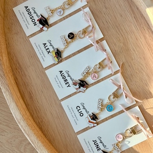 K&ouml;nnte beinhalten: Abschluss-Schl&uuml;sselanh&auml;nger mit goldfarbener Hardware, jeweils mit einem Charm mit einer Abschlusskappe, dem Jahr 2025 und einem Buchstaben. Die Schl&uuml;sselanh&auml;nger sind an Karten mit Namen wie Addison, Alex, Audrey, Clio und June befestigt und mit rosa B&auml;ndern gebunden.
