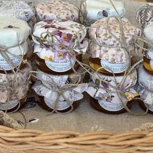 May include: Several small glass jars of honey are displayed in a woven basket. Each jar is covered with a fabric lid tied with twine. The lids feature floral and bee patterns. The jars have labels that read "Pure Honey, Tiptree, Essex, England."