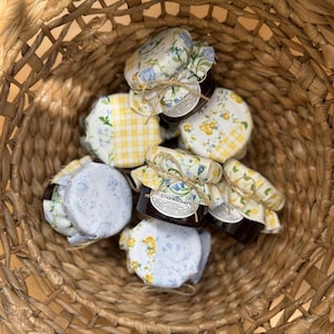 May include: A woven basket holds several small jars of preserves. Each jar is covered with a fabric lid tied with twine. The lids feature floral and checkered patterns in yellow, blue, and white. The jars contain a dark red preserve.