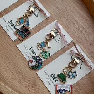 May include: Gold-tone keychains with camping-themed charms, including cameras, tents, and landscapes. Each keychain is attached to a card with a name and the text "Thanks for celebrating Hazel". The keychains are tied with pink ribbons.