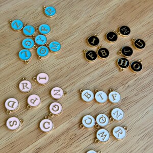 K&ouml;nnte beinhalten: Eine Sammlung goldfarbener Metallbuchstaben-Anh&auml;nger in verschiedenen Farben, darunter Hellblau, Schwarz, Rosa und Wei&szlig;. Jeder Anh&auml;nger zeigt einen anderen Buchstaben des Alphabets, ideal f&uuml;r die Schmuckherstellung oder personalisiertes Kunsthandwerk.