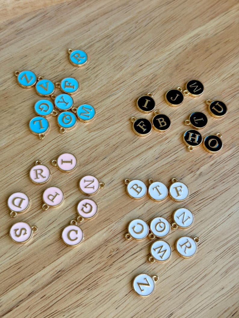 K&ouml;nnte beinhalten: Eine Sammlung goldfarbener Metallbuchstaben-Anh&auml;nger in verschiedenen Farben, darunter Hellblau, Schwarz, Rosa und Wei&szlig;. Jeder Anh&auml;nger zeigt einen anderen Buchstaben des Alphabets, ideal f&uuml;r die Schmuckherstellung oder personalisiertes Kunsthandwerk.