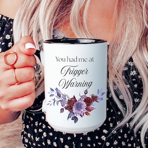 May include: White ceramic mug with a black rim, featuring the text "You had me at Trigger Warning" and a floral design in shades of purple and burgundy. The mug is held by a person wearing a black floral top and blue jeans.