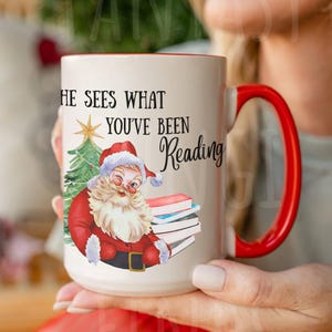 May include: A white ceramic mug with a red interior and handle. The mug features a Santa Claus illustration with the text "HE SEES WHAT YOU'VE BEEN Reading". The Santa illustration includes a Christmas tree and books.