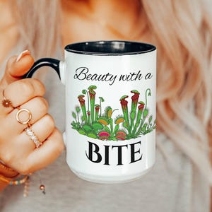May include: White ceramic mug with a black handle and rim, featuring the text "Beauty with a BITE" and a colorful illustration of carnivorous plants. The mug is held by a person.