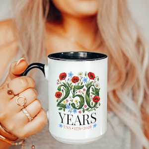 May include: White ceramic mug with a black handle and rim, featuring a floral design with red poppies, blue flowers, and daisies. The mug displays the text "250 YEARS USA 1776-2026".