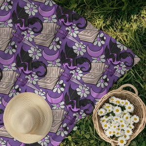 May include: A purple picnic blanket with a repeating pattern of black dragons, books, daggers, white daisies, and crescent moons. A straw hat and a basket of daisies sit on the grass next to the blanket.