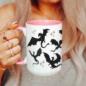 May include: White ceramic mug with a pink rim and handle. The mug features a black silhouette design of four dragons with gold stars.