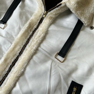 May include: Close-up of a white jacket with a cream-colored, fluffy collar and lining. The jacket features a gold zipper and black leather straps with gold buckles. The jacket is made of a smooth, white material.