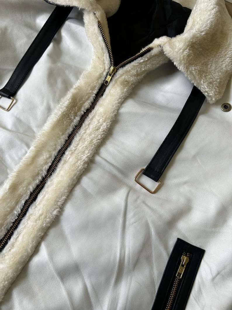 May include: Close-up of a white jacket with a cream-colored, fluffy collar and lining. The jacket features a gold zipper and black leather straps with gold buckles. The jacket is made of a smooth, white material.